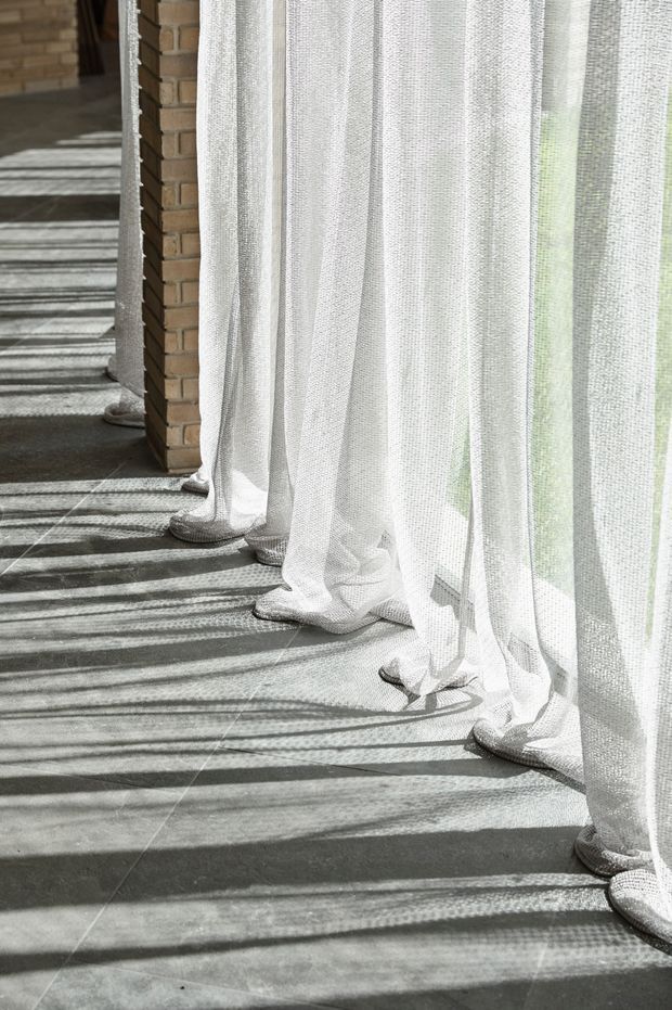 Close-up architectural interior detail showing sheer white floor-length curtains pooling on a tiled floor beside exposed warm-toned brick columns. Strong sunlight filters through the fabric, casting long striped shadows across the grey floor and creating a bright, textural atmosphere.