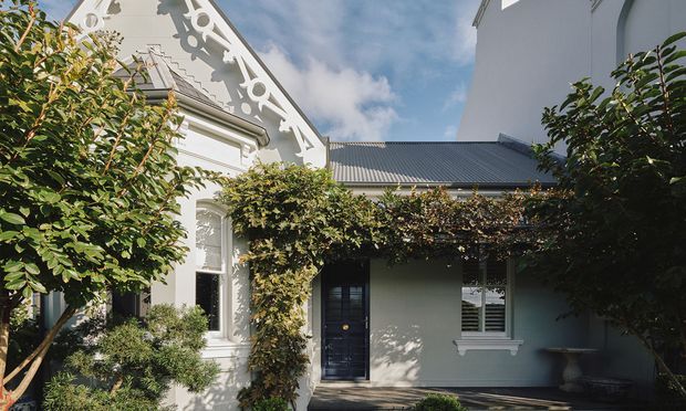 Exterior view of a traditional single-storey residence with ornate white gable detailing, decorative fretwork, rendered walls, a dark corrugated pitched roof, and a central entry shaded by established climbing greenery. The adjoining facade includes a dark glazed front door and white-trimmed windows, set within a leafy landscaped garden.