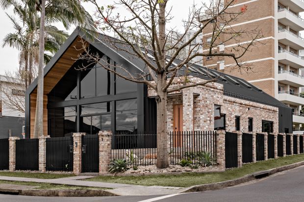 Street-level exterior view of a contemporary gabled building combining a dark metal-framed glazed frontage with a textured pale brick wing. The building has a steep pitched roof with dark roofing, exposed timber soffit detailing, black metal fencing, landscaped frontage, and a multi-storey apartment building visible behind it.