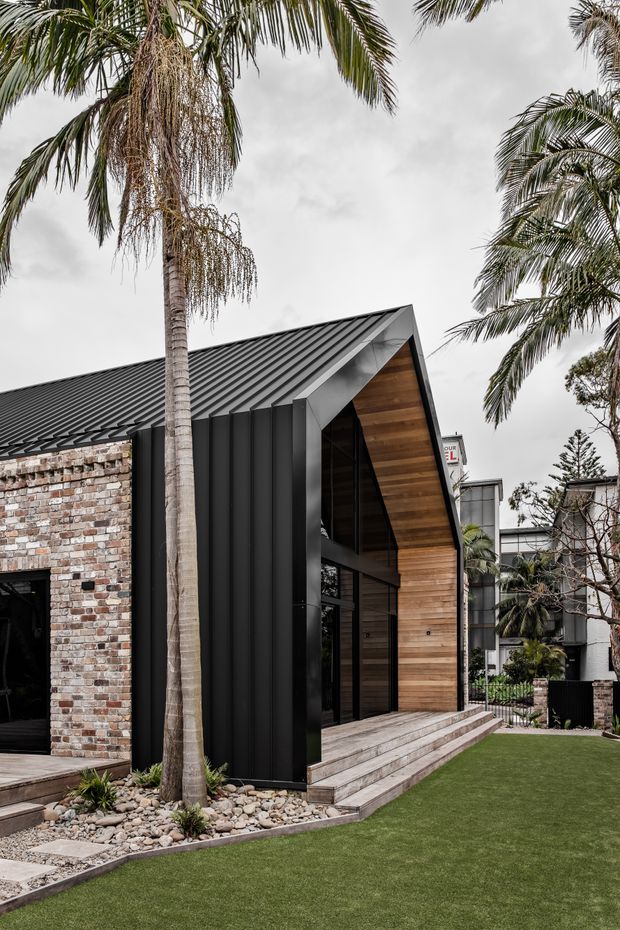 Exterior view of a contemporary gabled building with a steep dark metal roof, black vertical metal cladding, warm timber-lined gable and entry facade, and exposed mixed brick walls. A landscaped front yard with palms, pebble garden beds, paving and lawn surrounds the entrance.