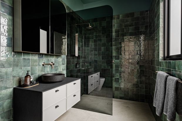 Moody contemporary bathroom with deep green glazed wall tiles, a black vanity with white drawers, a dark vessel basin, wall-mounted brushed metal tapware, large mirrored cabinetry, a walk-in shower, wall-hung toilet, and grey concrete-look flooring. Natural light enters through a black-framed window, while textured towels hang on a wall-mounted rail.