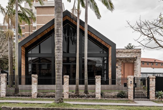 Street-facing view of a striking contemporary gabled building with a dark charcoal facade, extensive black-framed glazing, and warm timber lining beneath the deep triangular roof edges. A brick-and-metal perimeter fence, brick gate pillars, palm trees, and neighbouring urban buildings frame the property.