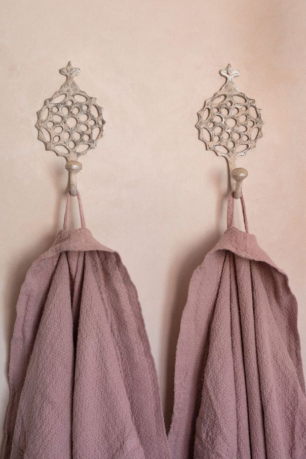 Close-up of two dusty-rose pink textured towels hanging from ornate silver-toned decorative wall hooks against a pale blush wall. The symmetrical arrangement emphasizes the intricate perforated metal hook design and soft fabric texture.