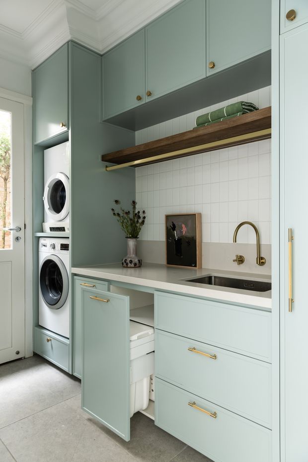 A compact, refined laundry room with pale sage-green cabinetry, stacked front-loading washer and dryer, a long light benchtop, integrated utility sink with brushed brass tapware, white square-tile splashback, and open dark timber shelving. Classic ceiling mouldings and brass hardware add a traditional touch, while a glass-panel exterior door brings in natural light.
