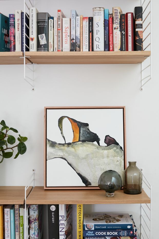A styled built-in or wall-mounted open shelving display against a white wall, with timber shelves supported by white metal brackets. The shelves hold an assortment of books, framed abstract artwork, glass vessels, ceramic vases, and a small leafy plant.