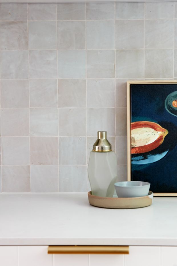 Close-up interior detail of a softly styled bathroom vanity or washroom area. Pale square ceramic wall tiles form the backsplash above a white benchtop, with light cabinetry below and a brass-toned horizontal handle. A small tray holding a decorative bottle and bowl sits beside a framed artwork leaning against the tiled wall.