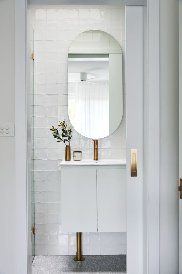 A compact, bright bathroom viewed through an open doorway, featuring glossy white rectangular wall tiles, a floating white vanity with integrated white basin, a tall arched-top mirror, and a brushed brass tap and wall switch. A small olive branch and ceramic vessel add a restrained natural accent. The floor is dark grey speckled tile, with white door and trim framing the space.