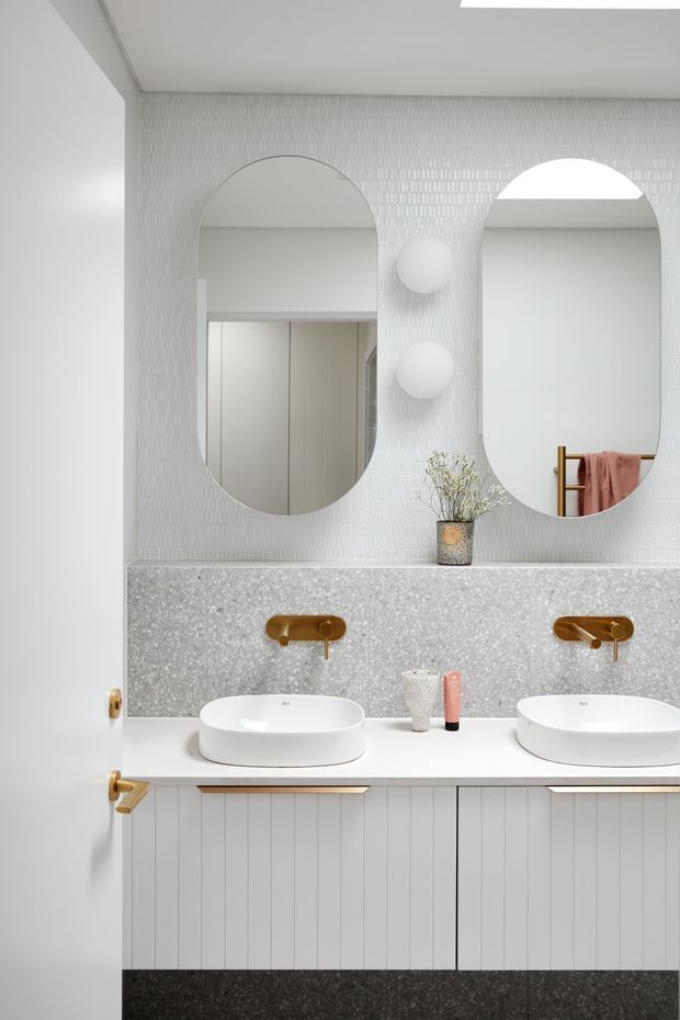 A bright, refined bathroom vanity featuring two white vessel basins on a long white benchtop, brushed brass wall-mounted taps, vertical-panel cabinetry, a speckled grey stone backsplash, and two large arched mirrors. The walls are covered in small white textured tiles, with globe wall lights and subtle decorative styling. A white door with brass hardware is visible at left.