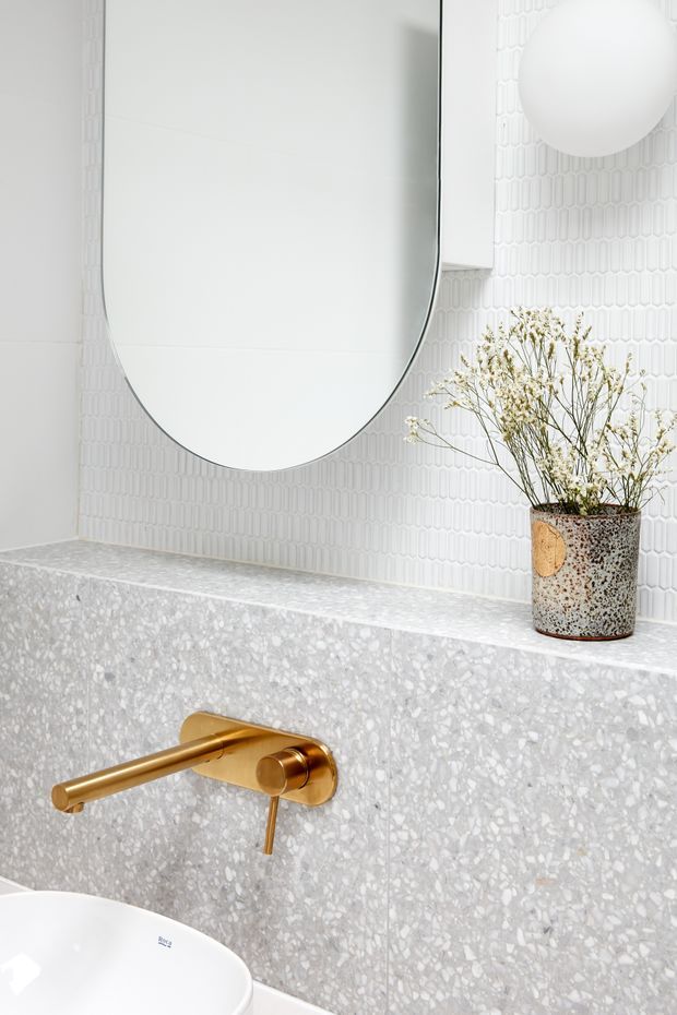 Close-up of a bright, refined bathroom vanity area featuring a large rounded-edge frameless mirror, a white vessel basin, brushed gold wall-mounted tapware, pale terrazzo wall and ledge surfaces, and a vertically patterned white tile backsplash. A speckled ceramic vase with delicate dried stems adds a natural decorative accent.