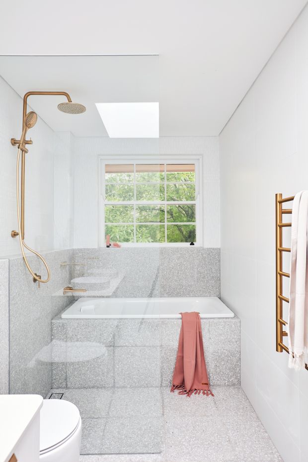 Bright contemporary bathroom featuring a built-in bathtub clad in speckled grey terrazzo, matching terrazzo wall and floor surfaces, white wall tiles, a white-framed window, skylight, brass rainfall shower and brass heated towel rail. A partial toilet and vanity are visible in the foreground, with greenery seen through the window.