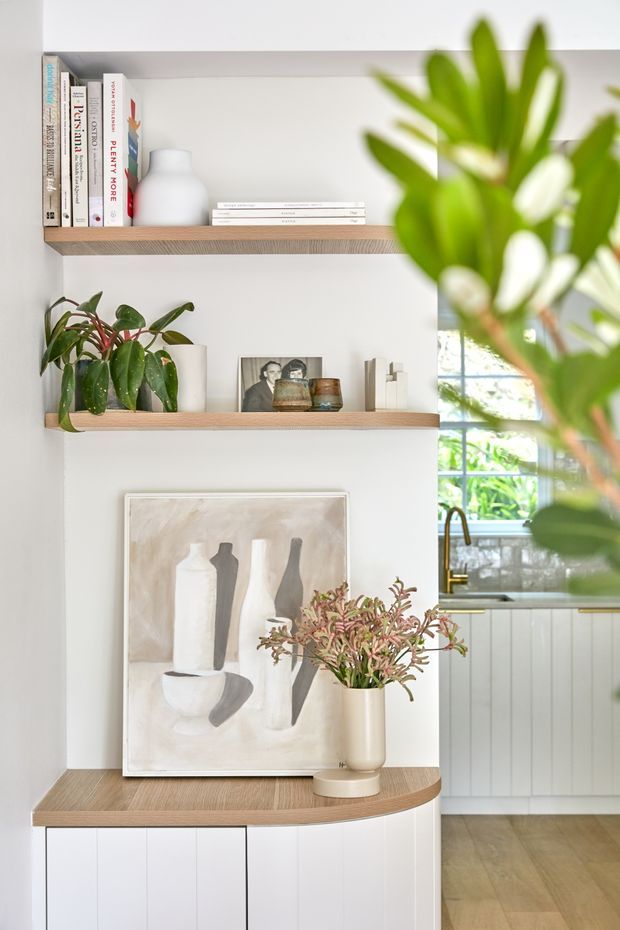 Bright, softly styled kitchen interior featuring open timber shelves with books, ceramics, plants and framed artwork, above a curved timber-topped white built-in cabinet. A partial kitchen is visible to the right with white cabinetry, a brass-toned tap, tiled splashback and a window overlooking greenery.
