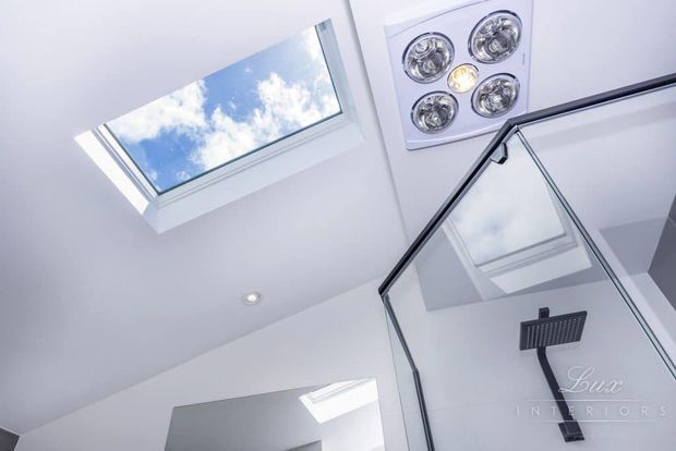 Upward-looking view of a bright contemporary bathroom shower area, featuring a frameless glass shower enclosure with a black shower fixture, white surfaces, a roof skylight, recessed ceiling lighting, and a ceiling-mounted ventilation or heat-light unit.