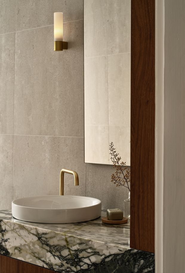 A refined contemporary bathroom vanity featuring a white vessel basin on a dramatic veined stone benchtop. Large grey-beige wall tiles, a tall frameless mirror, warm brass wall sconce and tapware, and a dark timber vertical panel create a warm, luxurious material palette.