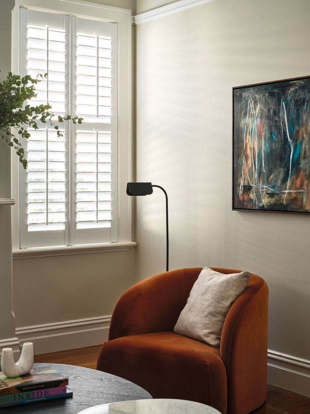 A warmly styled sitting area in a traditional room, featuring a rust-orange curved lounge chair with a linen cushion, a slim black floor lamp, a round dark stone coffee table, abstract artwork, white plantation shutters, and detailed wall mouldings. The space has dark timber flooring and soft neutral walls, with natural daylight creating a calm, refined atmosphere.