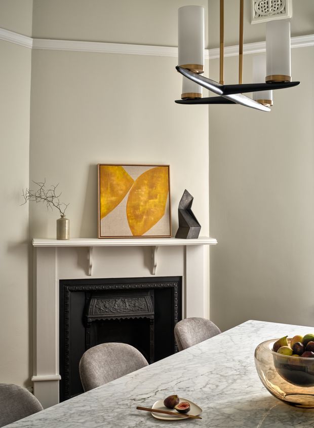 A refined dining area with a traditional decorative fireplace, ornate black fire surround, white mantel, and neutral plaster walls with detailed cornices. A sculptural contemporary pendant hangs above a veined marble dining table, accompanied by upholstered chairs. The mantel is styled with abstract artwork, a ceramic vase with branches, and a dark sculptural object, creating a blend of classic architectural detailing and contemporary furnishings.