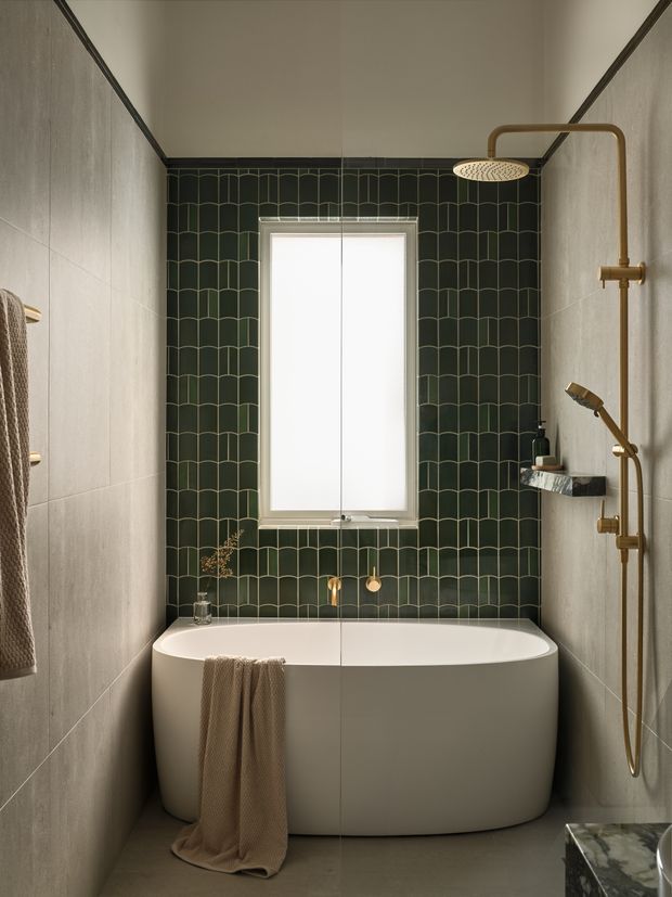 A compact, refined bathroom with a freestanding white bathtub positioned beneath a tall window. The rear wall is clad in dark green scalloped-pattern tiles with warm grout, while the side walls and floor use large pale stone-look tiles. Brushed brass shower fittings, a handheld shower, wall-mounted shelf, and minimal accessories create a warm contemporary look.