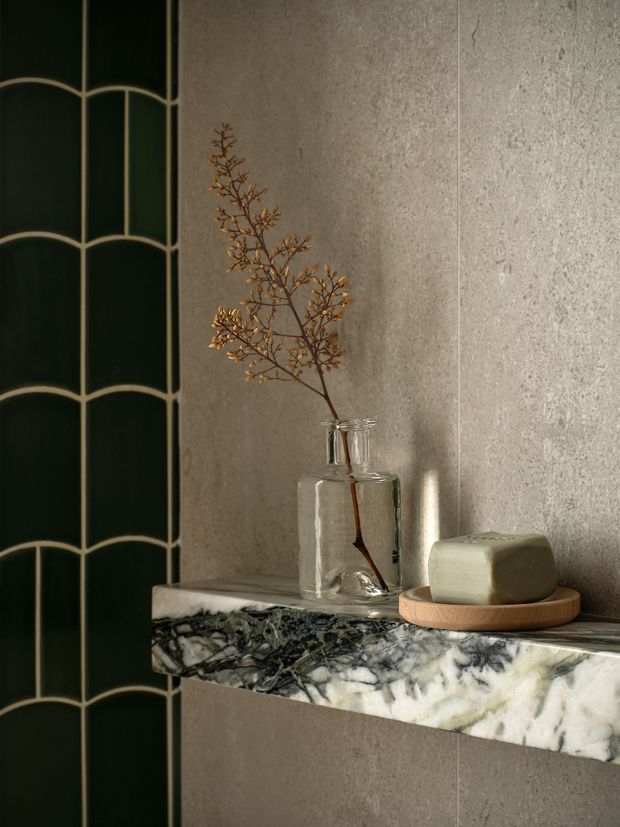 Close-up interior detail of a refined bathroom or washroom vignette. A dark green scalloped tile wall meets a textured grey stone-look wall, with a projecting veined marble shelf holding a clear glass vase with dried foliage and a muted green soap bar on a wooden dish. The composition is warm, moody and tactile, with strong emphasis on decorative wall finishes and natural stone.