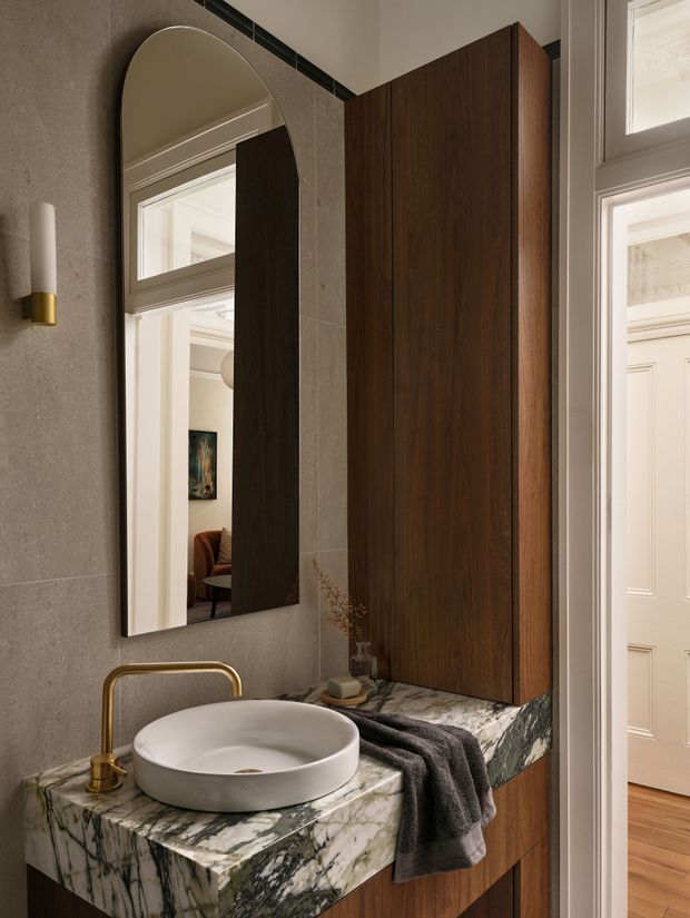 A refined bathroom vanity is shown in close-up, featuring a white vessel basin on a dramatic veined natural-stone benchtop, a brushed brass wall-mounted tap, dark timber cabinetry, and a large arched-edge mirror. Warm grey wall tiles, a slim dark accent tile band, and classic white interior doors visible beyond create a sophisticated traditional-contemporary atmosphere.