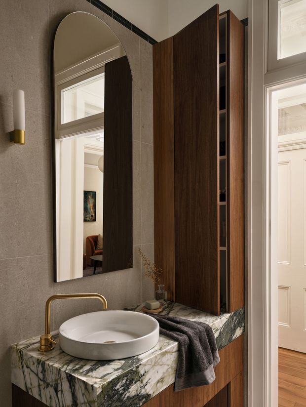 A refined bathroom vanity featuring a round vessel basin on a dramatic veined stone benchtop, brushed brass tapware, dark timber cabinetry, and a tall arched mirror. Grey textured wall tiles, warm integrated lighting, and a small wall sconce create a luxurious, moody atmosphere. A doorway and mirrored reflection reveal an adjoining interior space.
