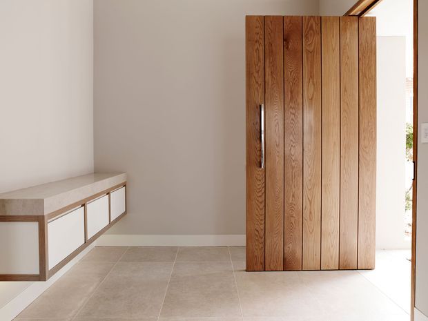 Minimalist entryway featuring a full-height pivot door clad in vertical natural timber boards, with a low built-in console on the left, pale tiled flooring, white walls, and bright natural light from the adjacent opening.