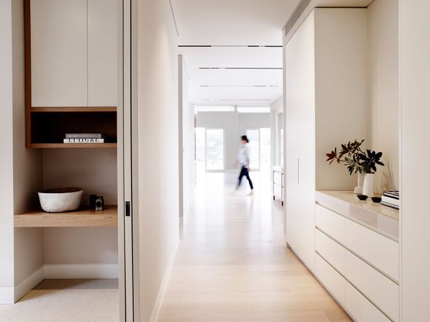 A bright, minimalist residential hallway with pale timber flooring and white walls. Built-in cabinetry and open shelving line both sides, with a concealed or flush door on the left and a low white storage unit topped with a vase on the right. The corridor leads toward a sunlit living area with large windows and a blurred person in the distance.