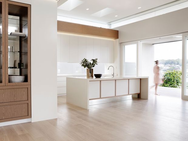 Bright, spacious contemporary kitchen with a large white island, integrated cabinetry, light timber flooring, and warm timber built-in display joinery. Full-height glazing opens toward an outdoor terrace, while a linear skylight and recessed ceiling lighting enhance the airy interior.