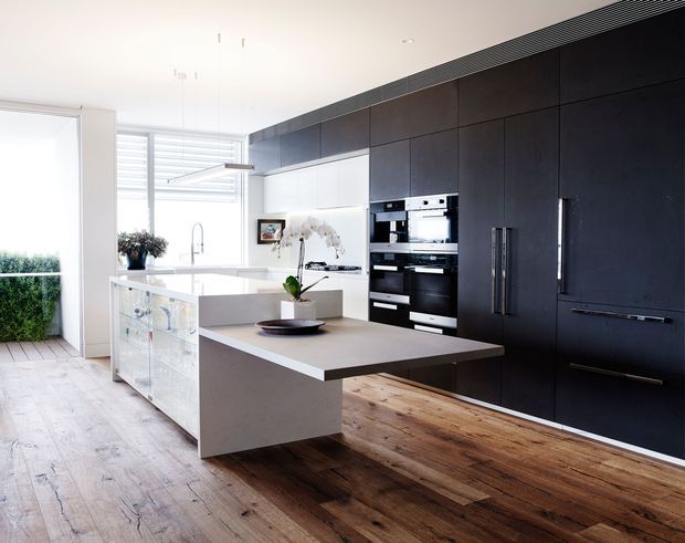 A bright, contemporary kitchen with a large central island, integrated black appliances, and a full wall of dark built-in cabinetry. The island has a pale stone-like benchtop and a white waterfall-style base, while the room features wide timber flooring, extensive glazing, and a clean, minimal fit-out.
