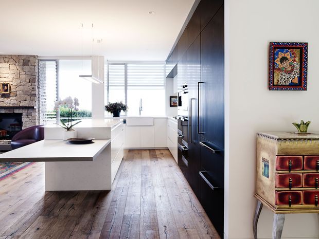 Bright contemporary open-plan kitchen and living area with white cabinetry, a large white island, dark full-height kitchen joinery, wide-plank timber flooring, and expansive louvred glazing. A rustic stone fireplace and seating area are visible to the left, while a colourful decorative cabinet and artwork add character on the right.