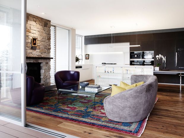 Bright open-plan living room with a contemporary white kitchen and island, dark built-in cabinetry, a stone fireplace, timber flooring, and a colorful patterned rug. The room features sculptural upholstered seating, glass coffee table, pendant lighting, and extensive sliding glass doors.