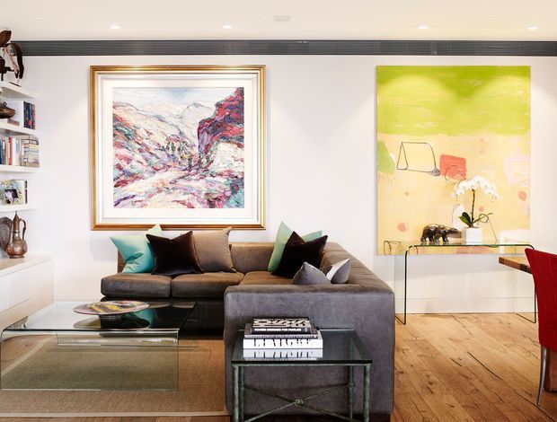 Contemporary living room with a large charcoal sectional sofa, colorful cushions, glass coffee table, framed artwork, white built-in cabinetry and shelving, timber flooring, and a red dining chair visible at the right edge. The room has a bright, curated gallery-like atmosphere with recessed ceiling lighting and a dark horizontal feature band near the ceiling.