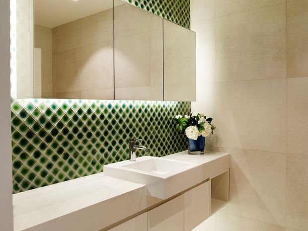 Contemporary bathroom vanity with a white integrated basin and wall-mounted cabinetry, set against large beige wall tiles. A striking green and white diamond-patterned tile backsplash runs beneath a wide mirrored cabinet, with a small blue vase and flowers adding colour.
