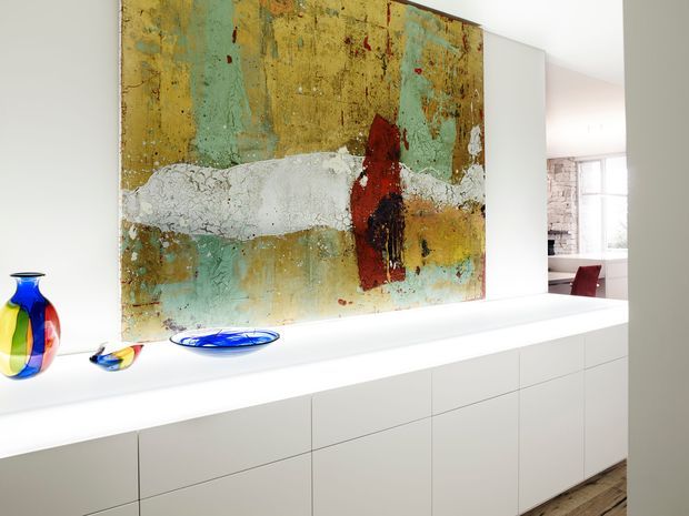 Bright contemporary interior featuring a long wall-mounted white built-in cabinet with handleless flat-front doors and a deep display ledge. A large abstract artwork in muted turquoise, ochre, white, and red dominates the wall above the joinery, complemented by colorful glass vessels. A dining area is partially visible beyond the open passage, with pale timber flooring and abundant natural light.