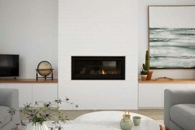 Bright contemporary living room featuring a white painted brick fireplace wall with a recessed gas fire, low white built-in cabinetry, a timber shelf, grey upholstered seating, a round light coffee table, and restrained botanical and art accents.