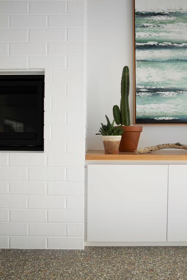 Close-up interior detail of a contemporary living room fireplace wall. A black inset fireplace is set into a white-painted brick surround, beside minimalist white built-in cabinetry with a light timber benchtop. Potted cacti, driftwood, and a framed abstract artwork add natural and coastal-inspired accents. The floor is finished in exposed terrazzo with multicoloured aggregate.