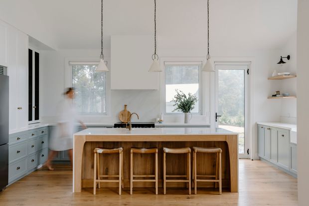 Bright, refined kitchen with a large white island featuring a warm timber base and four timber stools. The room includes white shaker-style cabinetry, pale timber flooring, marble-look benchtops and splashback, pendant lights, open shelving, and glazed doors and windows overlooking greenery. A softly blurred person is present near the cabinetry.