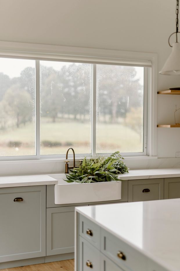 Bright, refined kitchen interior with muted sage-grey shaker cabinetry, white countertops, a large farmhouse sink beneath wide misted windows, brass-toned hardware, timber flooring, open shelving, and a white pendant light. A central island occupies the foreground, while soft greenery and a pastoral landscape are visible through the glazing.