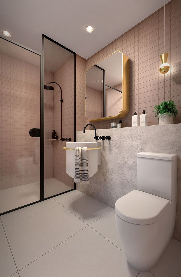 A compact contemporary bathroom featuring blush-pink small-format tiled walls, pale large-format floor tiles, a frameless black-framed glass shower enclosure, a floating concrete-look vanity with integrated basin, black wall-mounted tapware, a gold-edged geometric mirror, and a white toilet. Warm globe wall lighting and a small potted plant add refined decorative accents.
