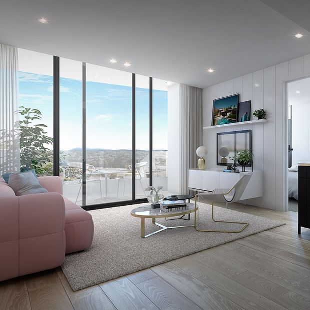 Bright contemporary living room with a large pink sectional sofa, pale textured area rug, glass and metal coffee table, sculptural lounge chair, and white built-in media and display cabinetry. Full-height black-framed glazing opens to a balcony and expansive city view, while sheer curtains, indoor plants, and recessed ceiling lights create an airy, polished apartment interior.