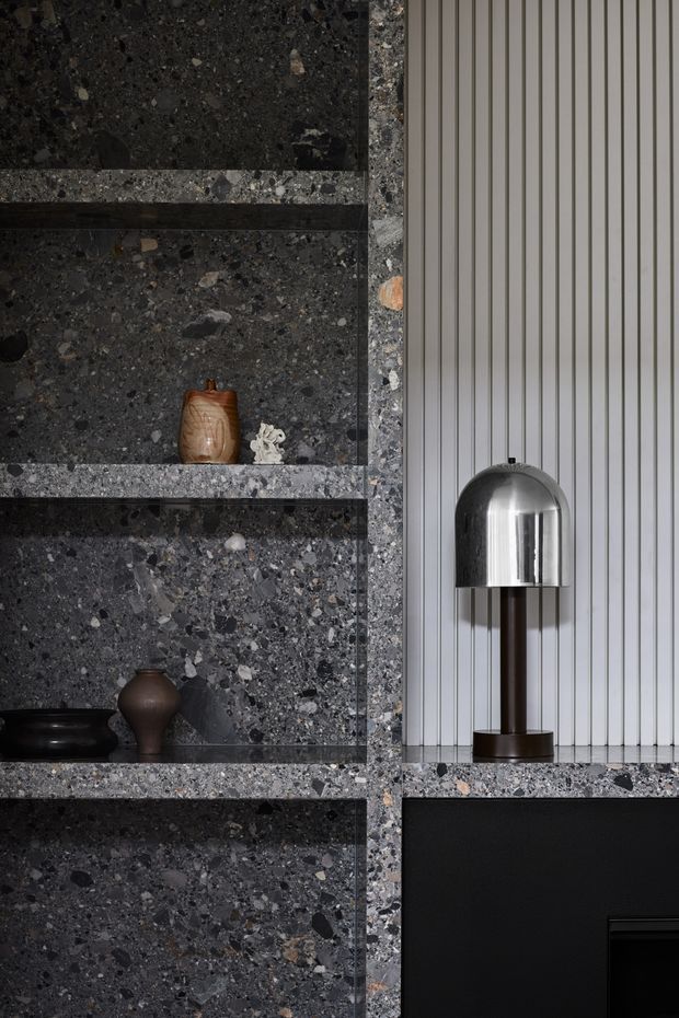Close-up of a refined contemporary interior feature wall combining dark, heavily speckled terrazzo or aggregate stone shelving with integrated black horizontal shelves and cabinetry. A vertically fluted light-grey wall panel sits beside the shelving, supporting a polished chrome dome table lamp. Small sculptural ceramic objects add restrained decorative detail.