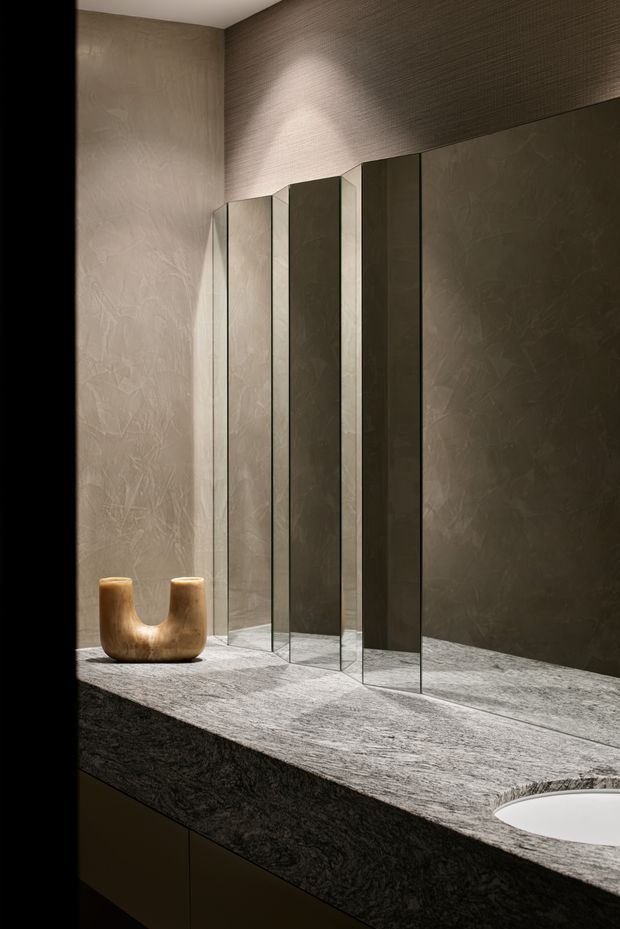 A moody, refined bathroom vanity viewed close-up. The space features a long dark stone vanity benchtop with an integrated white basin, frameless mirrored panels, dark textured wall finishes, and subtle concealed lighting. A small sculptural timber vessel provides a warm natural accent.