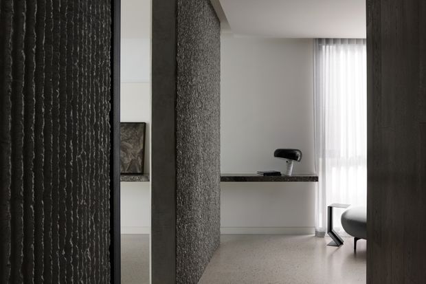 A refined contemporary interior viewed through a dark textured partition, showing a softly lit living or lounge area with a curved chair, slim black side table, built-in low shelf, artwork, and sheer floor-to-ceiling curtains. The space features neutral grey and off-white tones, textured feature surfaces, and a restrained minimalist composition.