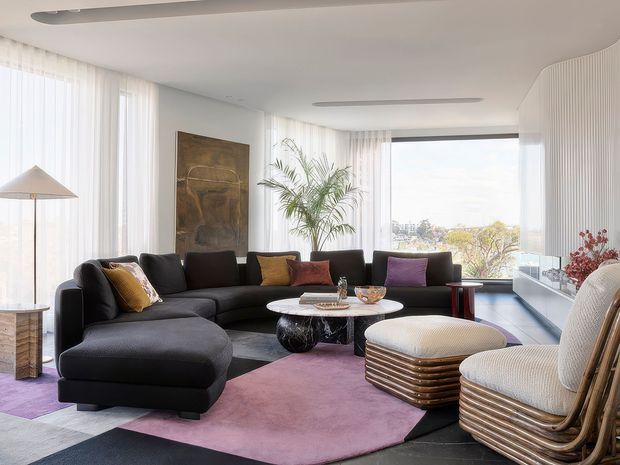 Bright, spacious contemporary living room with floor-to-ceiling sheer curtains and expansive glazing. A large black sectional sofa anchors the room, complemented by sculptural coffee tables, layered geometric rugs in mauve, grey and black, rounded upholstered lounge chairs, and a tall indoor palm. Curved white walls and linear ceiling lights create a refined, softly organic interior with city and greenery views beyond the windows.