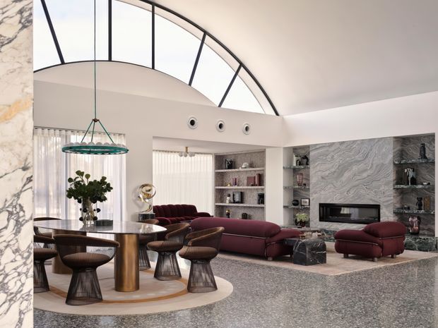 A refined open-plan living and dining area with a dramatic curved clerestory window and vaulted ceiling. The room features a dark stone or terrazzo floor, a round dining table with sculptural upholstered chairs, a suspended circular pendant, burgundy seating, built-in shelving, and a fireplace set within a richly veined marble feature wall. The palette is neutral with deep burgundy accents and an upscale contemporary feel.