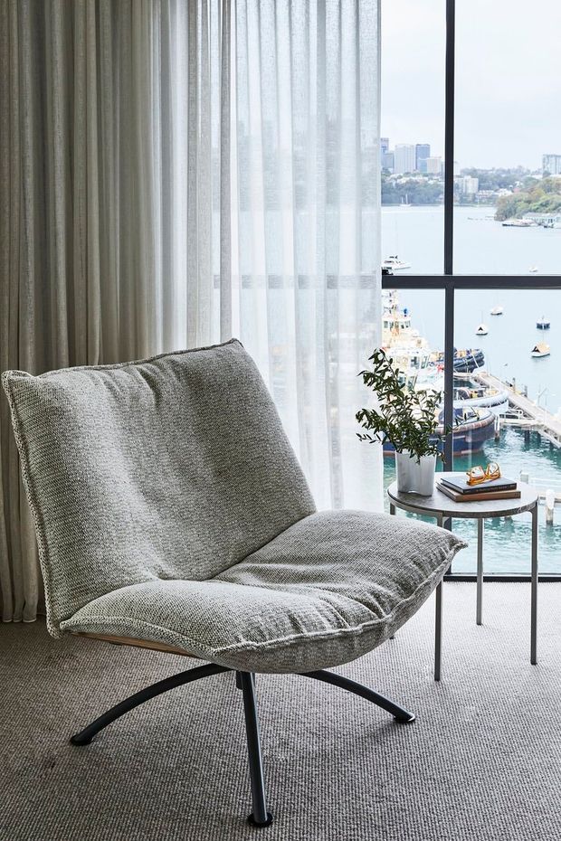 A softly lit waterfront-facing interior corner with a sculptural upholstered lounge chair in textured grey fabric, positioned beside a slim round side table with a potted plant and books. Floor-to-ceiling sheer curtains and dark-framed glazing reveal a marina and city waterfront beyond. The palette is muted and refined, with carpeted flooring and layered window treatments.