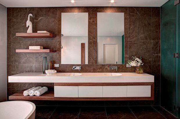A sophisticated contemporary bathroom featuring a long double vanity with a pale stone benchtop, white integrated basins, and dark timber drawer fronts. Large dark brown marble-look wall and floor tiles create a dramatic backdrop, complemented by two frameless mirrors, open timber display shelves, recessed ceiling lights, and a freestanding bath partially visible in the foreground. A dark green door or partition adds a contrasting accent.