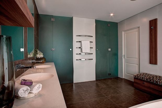 A spacious contemporary bathroom with a long pale stone double vanity, two vessel basins, large wall mirror, dark tiled flooring, teal glass shower or enclosure doors, chrome heated towel rails, and a paneled white door. A timber ceiling or wall feature and upholstered bench add warmth to the otherwise refined interior.