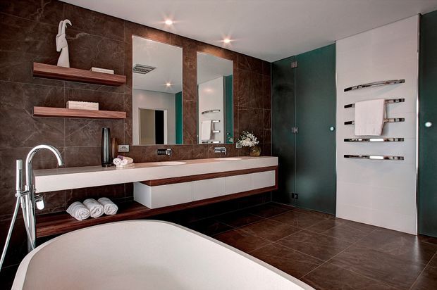 Contemporary bathroom with a freestanding bathtub in the foreground, a long double vanity with wall-mounted mirrors, dark brown tiled walls and floor, floating wood-and-white cabinetry, open display shelves, and a glass-enclosed shower area. Chrome fixtures and heated towel rails add polished functional details.