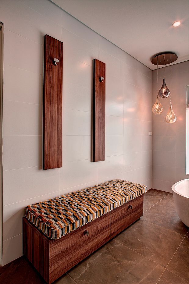 A refined bathroom interior featuring large light beige wall tiles, darker brown floor tiles, a freestanding bathtub, and a long built-in timber bench with drawers and a patterned upholstered seat. Two tall timber wall panels with small hooks or fittings are mounted above the bench, while a cluster of decorative pendant lights hangs from a circular ceiling plate.