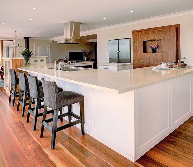 Bright, spacious open-plan kitchen dominated by a large white island with a pale stone benchtop and seating for five. The room features white paneled cabinetry, timber flooring, stainless-steel appliances and rangehood, recessed ceiling lighting, and a timber feature wall with a recessed display niche. A dining or living area is partially visible beyond the kitchen.