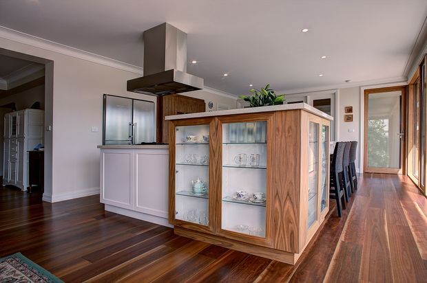 Bright open-plan kitchen and dining area with a prominent custom timber display cabinet or island featuring glass-fronted shelving, set on dark polished timber flooring. The kitchen includes a pale island, stainless-steel rangehood, dark timber cabinetry, recessed ceiling lights, and a row of upholstered bar stools beside large glazed doors and windows.